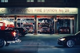 A city fire station with a sign indicating 'City of Houston Fire Station No. 28'. Several fire trucks and emergency vehicles are visible inside the garage bays, along with holiday lights decorating the station. A few cars are parked or moving in front of the station, and a person is seen walking nearby.