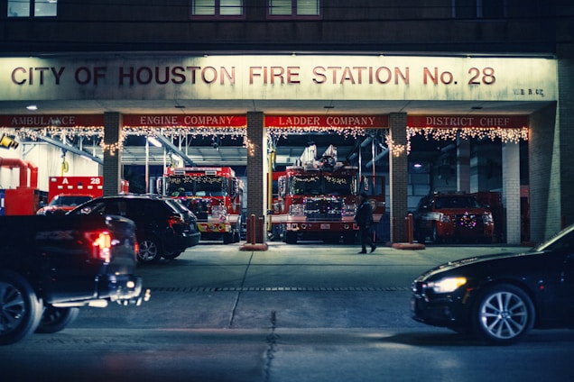 A city fire station with a sign indicating 'City of Houston Fire Station No. 28'. Several fire trucks and emergency vehicles are visible inside the garage bays, along with holiday lights decorating the station. A few cars are parked or moving in front of the station, and a person is seen walking nearby.