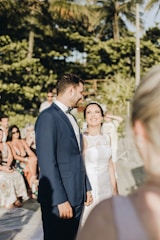 A couple dressed in wedding attire stands smiling at each other in an outdoor setting, surrounded by seated guests. The background features lush green foliage and partially visible palm trees under sunny weather.