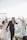 A joyful wedding scene on a beach with a bride and groom walking hand in hand. The groom is dressed in a navy blue suit and the bride in a white gown, holding a bouquet. A woman in a light blue dress is cheering with her hands up. There are white floral decorations and the ocean is visible in the background, along with some chairs and a photographer capturing the moment.
