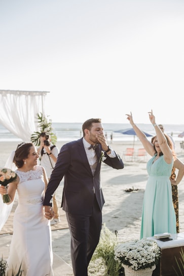 A joyful wedding scene on a beach with a bride and groom walking hand in hand. The groom is dressed in a navy blue suit and the bride in a white gown, holding a bouquet. A woman in a light blue dress is cheering with her hands up. There are white floral decorations and the ocean is visible in the background, along with some chairs and a photographer capturing the moment.