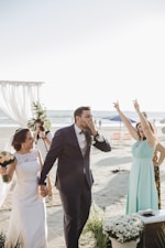 A joyful wedding scene on a beach with a bride and groom walking hand in hand. The groom is dressed in a navy blue suit and the bride in a white gown, holding a bouquet. A woman in a light blue dress is cheering with her hands up. There are white floral decorations and the ocean is visible in the background, along with some chairs and a photographer capturing the moment.