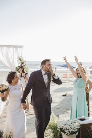 A joyful wedding scene on a beach with a bride and groom walking hand in hand. The groom is dressed in a navy blue suit and the bride in a white gown, holding a bouquet. A woman in a light blue dress is cheering with her hands up. There are white floral decorations and the ocean is visible in the background, along with some chairs and a photographer capturing the moment.
