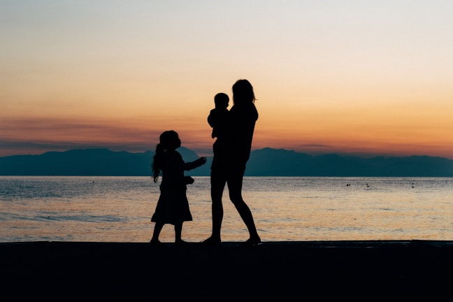 A silhouette of a woman holding a child and a girl standing next to her, set against the backdrop of a sunset over a calm sea. The sky is painted in warm orange and yellow hues, with a range of mountains in the distance.