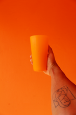 A hand holding an orange cup against an orange background, with a tattoo visible on the arm holding the cup.