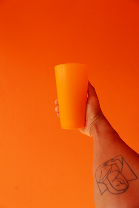 A hand holding an orange cup against an orange background, with a tattoo visible on the arm holding the cup.