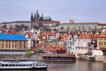A river scene featuring a boat labeled 'Prague Boats' passing in front of a historical cityscape. The background includes a row of colorful buildings with red-tiled roofs, and prominent architectural structures including a large castle-like complex with spires and green domes.