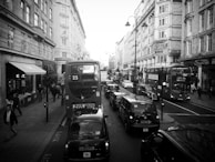 A busy city street with several double-decker buses and black cabs in traffic. People are walking on the sidewalks past various storefronts and restaurants. Historical buildings with ornate architecture line the street, and there is a prominent sign for the Strand Palace Hotel.