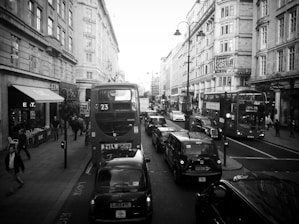 A busy city street with several double-decker buses and black cabs in traffic. People are walking on the sidewalks past various storefronts and restaurants. Historical buildings with ornate architecture line the street, and there is a prominent sign for the Strand Palace Hotel.