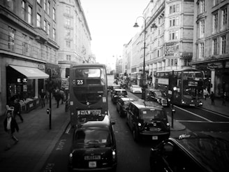 A busy city street with several double-decker buses and black cabs in traffic. People are walking on the sidewalks past various storefronts and restaurants. Historical buildings with ornate architecture line the street, and there is a prominent sign for the Strand Palace Hotel.