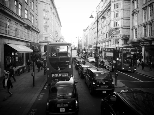 A busy city street with several double-decker buses and black cabs in traffic. People are walking on the sidewalks past various storefronts and restaurants. Historical buildings with ornate architecture line the street, and there is a prominent sign for the Strand Palace Hotel.