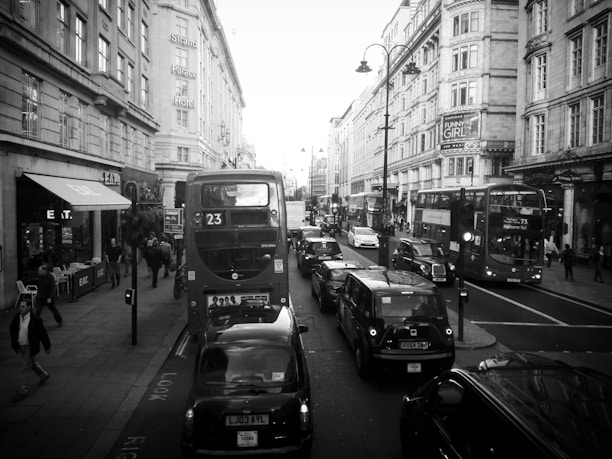 A busy city street with several double-decker buses and black cabs in traffic. People are walking on the sidewalks past various storefronts and restaurants. Historical buildings with ornate architecture line the street, and there is a prominent sign for the Strand Palace Hotel.