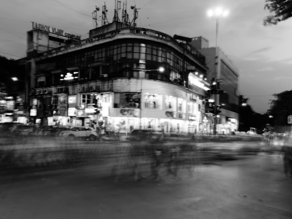 A busy street scene at dusk in a city with blurred motion, likely from cars and people, creating a sense of movement. A multi-story building with numerous windows and lit signs is prominent in the image, with antennas on the roof. Streetlights provide illumination, contrasting with the dimming sky.