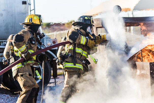 Two firefighters in full gear are actively extinguishing a fire using a hose, with flames and smoke visible around them. They are wearing helmets and breathing apparatus, working in close coordination to control the blaze.