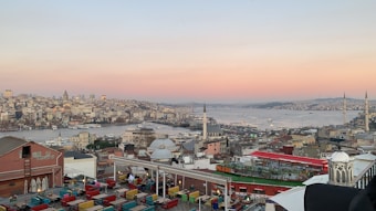 A panoramic view of a city with a large body of water in the center. The skyline includes a mix of historic and modern buildings, with several minarets visible. The foreground features brightly colored seating on a rooftop terrace overlooking the city. The sky displays a gentle gradient from pink to blue, indicating either sunrise or sunset.