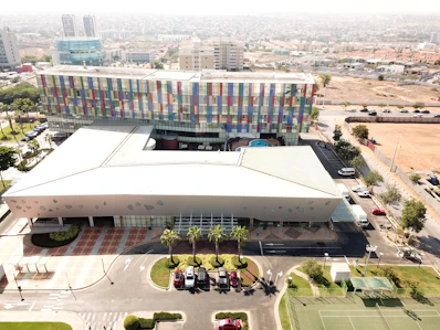 An aerial view of a large modern building complex with a colorful facade, featuring panels in various shades of red, blue, and green. The complex is surrounded by roads and includes a few parked cars and well-manicured landscaping, such as palm trees and grassy areas. There are tennis courts visible in the foreground and a residential area in the background.