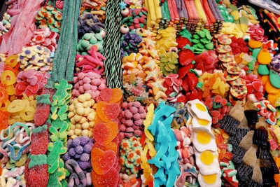 A vibrant assortment of colorful candies, including gummy bears, jelly beans, candy strips, sugared fruit slices, and cola bottles. The candies are arranged in a visually appealing manner, with a variety of shapes, sizes, and textures.