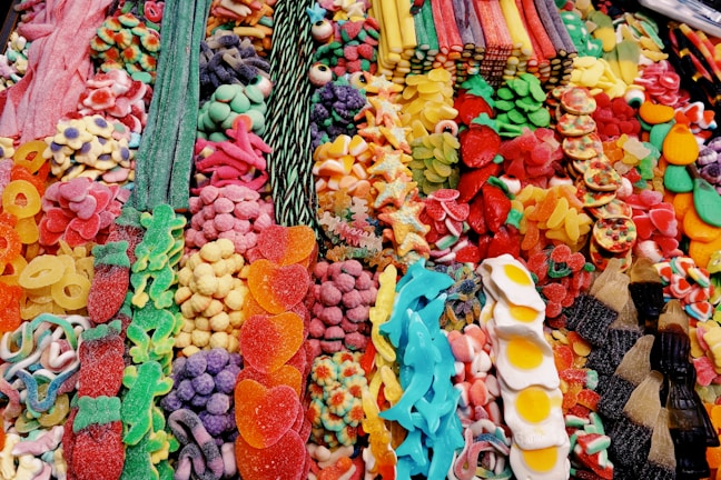 A vibrant assortment of colorful candies, including gummy bears, jelly beans, candy strips, sugared fruit slices, and cola bottles. The candies are arranged in a visually appealing manner, with a variety of shapes, sizes, and textures.