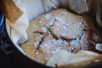 A freshly baked bread loaf with a golden brown crust, resting on parchment paper. The surface is dusted with flour and features a rustic, cracked texture.