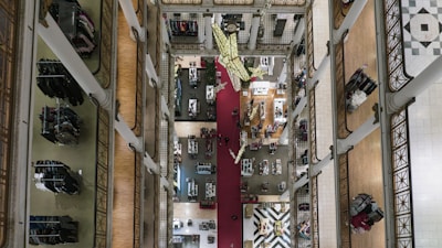 Aerial view of a multi-story department store with tall white pillars, intricate railings, and clothing racks neatly organized along the corridors. A decorative red carpet runs down the center, with a geometric pattern floor visible on the lower level. Shoppers can be seen browsing the goods.
