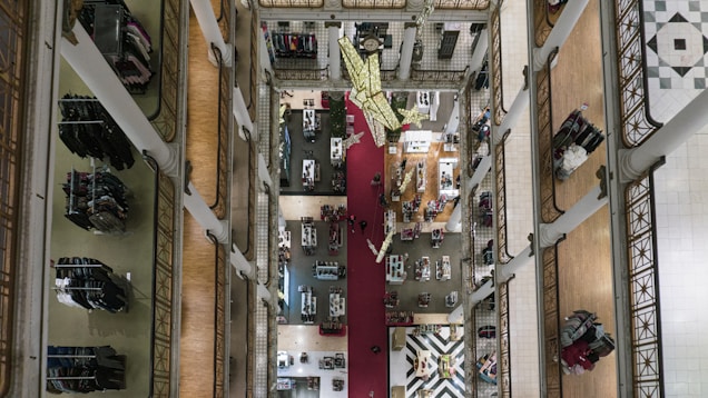 Aerial view of a multi-story department store with tall white pillars, intricate railings, and clothing racks neatly organized along the corridors. A decorative red carpet runs down the center, with a geometric pattern floor visible on the lower level. Shoppers can be seen browsing the goods.