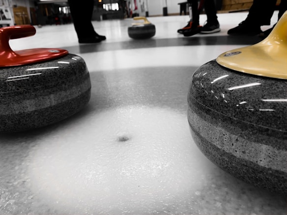 Close-up view of curling stones on a curling rink, with three stones visible—one with a red handle and another with a yellow handle. The ice surface is textured and reflects light. In the background, the legs and feet of several people can be seen, indicating participation in the sport.