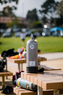 A scuba diving tank stands upright on a bench in an outdoor setting with grass and trees in the background. Another tank lies horizontally on the bench with various diving gear scattered nearby.