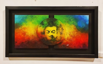 A colorful painting features a serene face within a circular cutout at the center of a vibrant background blending rainbow colors including red, green, blue, and yellow. The texture appears tactile with swirling patterns and abstract designs.