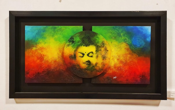 A colorful painting features a serene face within a circular cutout at the center of a vibrant background blending rainbow colors including red, green, blue, and yellow. The texture appears tactile with swirling patterns and abstract designs.