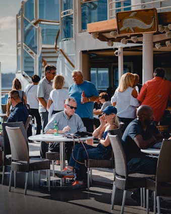 A group of people are socializing on an outdoor deck area, possibly on a cruise ship or similar setting. Several individuals are seated at round tables with wicker chairs, engaging in conversation. The background features a modern structure with glass and metal elements, and a wooden sign with a dolphin logo. Some people are standing, while others are seated, creating a lively and casual atmosphere.