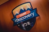 A decorative sign prominently features the words 'Camp Discovery' with stylized mountain peaks depicted above. The sign is set against a wooden background, and the earth is included in the design where the letter 'o' is in Discovery. The colors are bold and vibrant, consisting of blues, oranges, and whites.