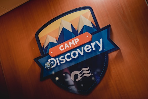 A decorative sign prominently features the words 'Camp Discovery' with stylized mountain peaks depicted above. The sign is set against a wooden background, and the earth is included in the design where the letter 'o' is in Discovery. The colors are bold and vibrant, consisting of blues, oranges, and whites.