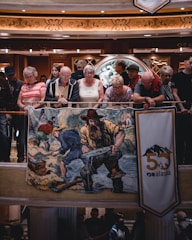 A group of people, mostly elderly, are standing and leaning on a railing indoors. They are observing something below them. In front of them, there is a large colorful banner featuring a historic scene of gold miners at a river. There is also a vertical banner with the text 'Alaska' and the number '50'. The environment appears to be well-lit and the architectural details include intricate designs and warm lighting.