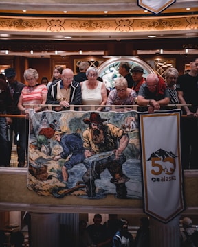 A group of people, mostly elderly, are standing and leaning on a railing indoors. They are observing something below them. In front of them, there is a large colorful banner featuring a historic scene of gold miners at a river. There is also a vertical banner with the text 'Alaska' and the number '50'. The environment appears to be well-lit and the architectural details include intricate designs and warm lighting.