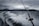 A fishing rod is angled toward the water as a boat speeds along, creating a trail of white spray on a dark, choppy sea. In the background, a mountain range looms under a cloudy, overcast sky, lending a dramatic atmosphere.