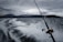 A fishing rod is angled toward the water as a boat speeds along, creating a trail of white spray on a dark, choppy sea. In the background, a mountain range looms under a cloudy, overcast sky, lending a dramatic atmosphere.