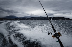 A fishing rod is angled toward the water as a boat speeds along, creating a trail of white spray on a dark, choppy sea. In the background, a mountain range looms under a cloudy, overcast sky, lending a dramatic atmosphere.