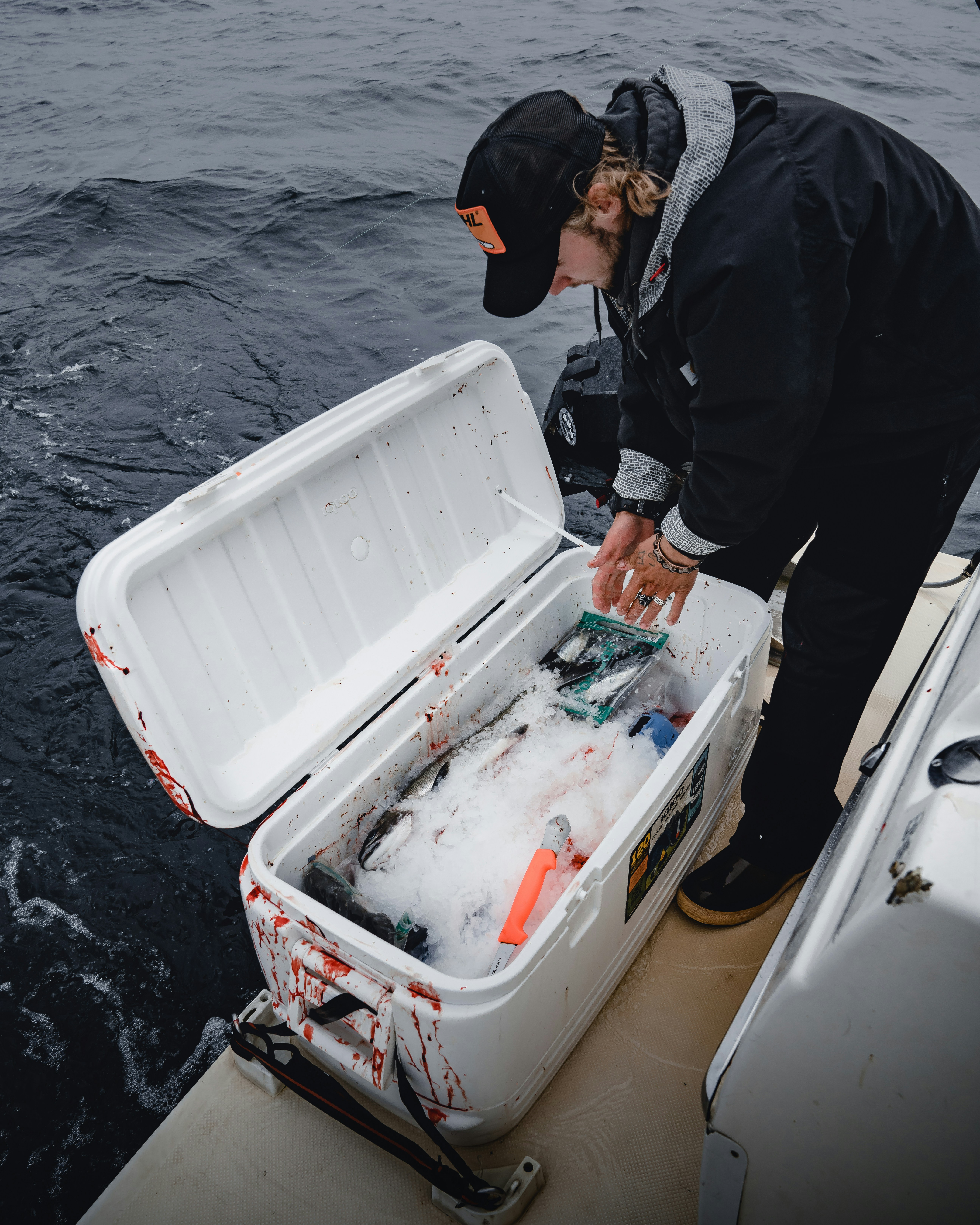 Angler reaching into an open white cooler on a boat, fresh fish packed in crushed ice, dark water alongside