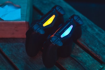 Two gaming controllers with illuminated light bars, one displaying a yellow glow and the other a blue glow, are placed on a wooden surface. To the left, there is a part of a wooden crate with a white paper object on it.