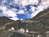 A road curves through a mountainous landscape with a hillside adorned by white chortens and a cluster of building structures. The sky above is filled with dramatic clouds against patches of blue.