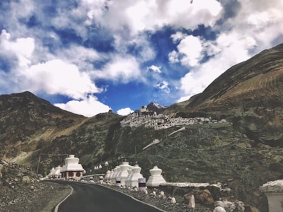 A road curves through a mountainous landscape with a hillside adorned by white chortens and a cluster of building structures. The sky above is filled with dramatic clouds against patches of blue.