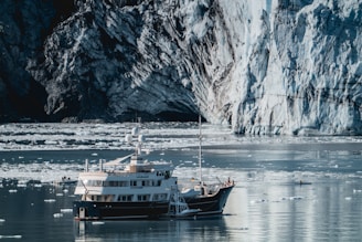 A large yacht is anchored in icy waters surrounded by small icebergs. The backdrop features a massive glacier with rugged textures and blue-white hues. A small inflatable boat is nearby, and the water reflects the cold environment.