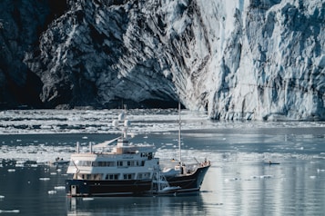 A large yacht is anchored in icy waters surrounded by small icebergs. The backdrop features a massive glacier with rugged textures and blue-white hues. A small inflatable boat is nearby, and the water reflects the cold environment.