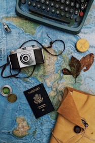 A vintage camera, a typewriter, a passport, and several travel-related items are arranged on an old world map. The collection includes a compass, a bottle with a small ship, a yellow envelope, and dried leaves, evoking a sense of nostalgia and adventure.