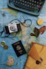 A vintage camera, a typewriter, a passport, and several travel-related items are arranged on an old world map. The collection includes a compass, a bottle with a small ship, a yellow envelope, and dried leaves, evoking a sense of nostalgia and adventure.