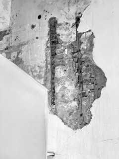 A section of a wall with significant damage, showing exposed bricks and rough concrete. There are various marks and holes visible on the wall. The paint appears worn and peeling in several areas.
