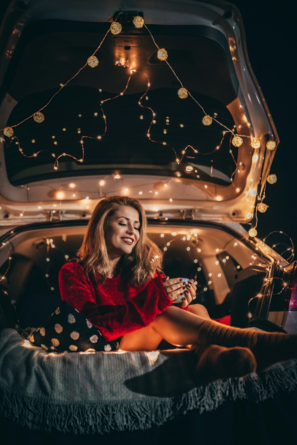 smiling woman wearing red crew-neck sweatshirt sitting at the back of vehicle while holding phone surrounded with lighted string lights