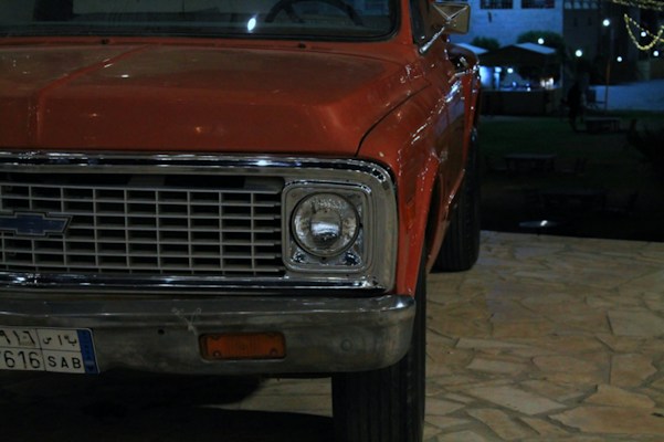 The image features the front portion of a vintage car with a rusty red exterior, prominently displaying a classic grille and a headlight. The scene is set on a tiled patio during the evening, with dim ambient lighting and a building in the background.
