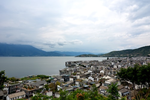 A scenic view of a lakeside town with houses clustered along the shoreline. The calm water extends to the distant hills and mountain range under a cloudy sky.