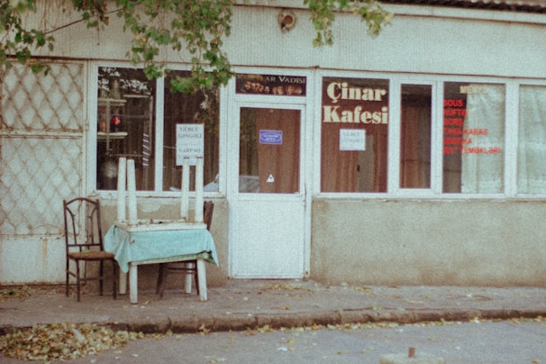 An exterior view of a small, slightly weathered café with signs on the windows and door, situated on a quiet street. The café is named 'Çınar Kafesi'. A table with chairs and a light blue tablecloth sits outside, with upside-down chairs placed on top. A few leafy branches hang down from above.